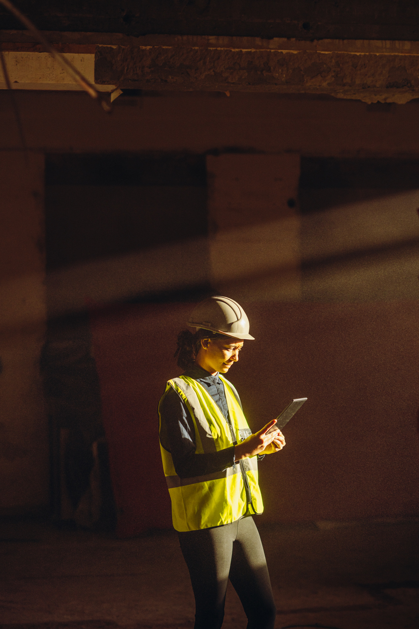 Female Worker indoors on Construction Site with sun beam falling on dust with PPE on tablet.